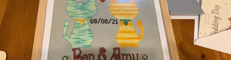 A photograph of a rectangular piece of light grey cross-stitch Aida in a light wood frame. The design shows two cat silhouettes: one is variegated green with an orange collar and a gold tag, the other variegated yellow with a green collar and a green tag. Between them is a red heart and the date 08/08/21. Beneath are the words "Ben & Amy" in a red font stylised with hearts in the capital letters, plus two pairs of interlocked green and gold wedding rings.