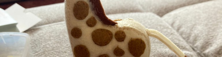 A photograph of a small felt giraffe. It's more realistic in appearance and is yellow with brown spots and a brown mane.