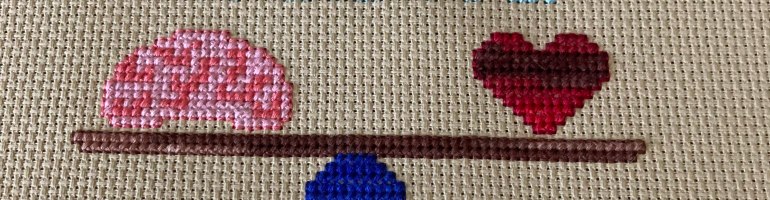 A photograph of a cross-stitch. A variegated brown seesaw sits on top of a dark blue base. On one side is a brain in various shades of pink and on the other is a variegated red heart. Both are perfectly balanced. Above and below it are the words "Find Your Wise Mind" in light blue thread. The words are not balanced at all.