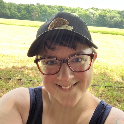 Penny's author profile picture. In it, Penny is looking at the camera and smiling. They are a short-haired white person with red glasses and a black baseball cap with a hedgehog on it.